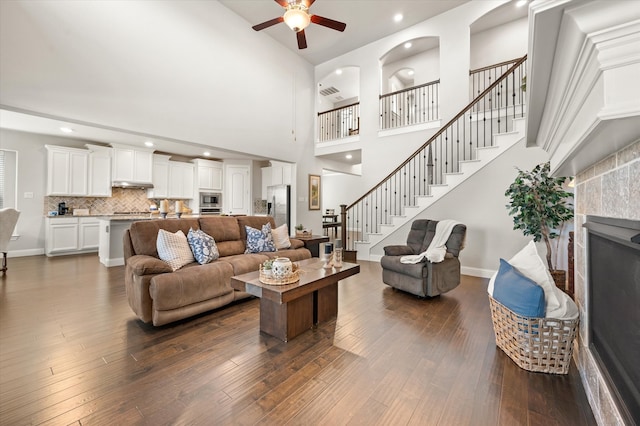 living room with a towering ceiling, ceiling fan, dark wood-type flooring, and a tiled fireplace