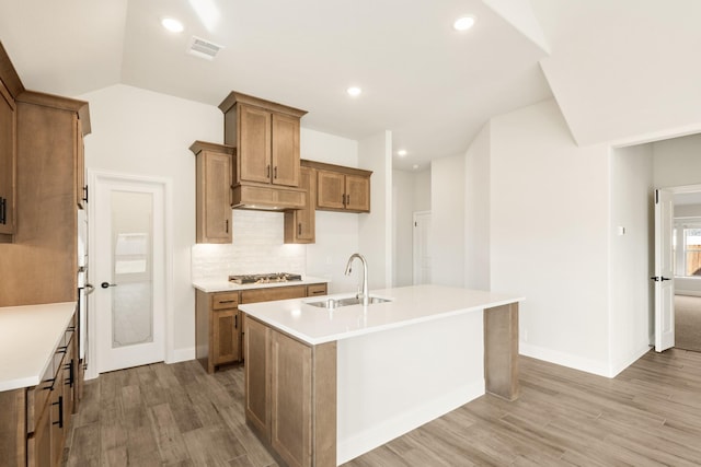 kitchen with brown cabinetry, an island with sink, a sink, stainless steel gas stovetop, and backsplash