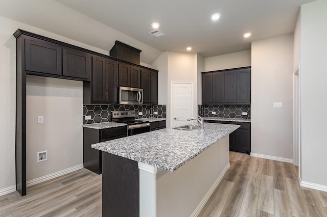 kitchen featuring appliances with stainless steel finishes, light wood-type flooring, tasteful backsplash, light stone counters, and an island with sink