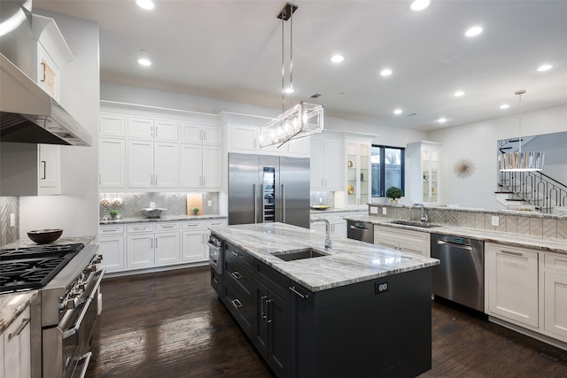 kitchen with premium appliances, wall chimney range hood, a center island with sink, white cabinets, and hanging light fixtures