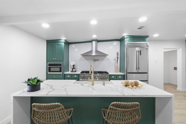 kitchen with high end appliances, light stone countertops, wall chimney range hood, green cabinets, and light wood-type flooring