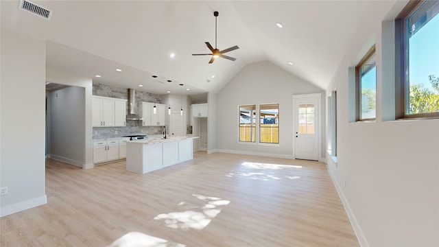 kitchen with wall chimney exhaust hood, an island with sink, decorative light fixtures, white cabinets, and light wood-type flooring