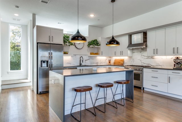 kitchen with wall chimney exhaust hood, hanging light fixtures, stainless steel appliances, a kitchen island, and hardwood / wood-style flooring