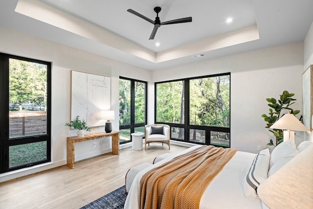 bedroom with ceiling fan, light hardwood / wood-style floors, a raised ceiling, and multiple windows