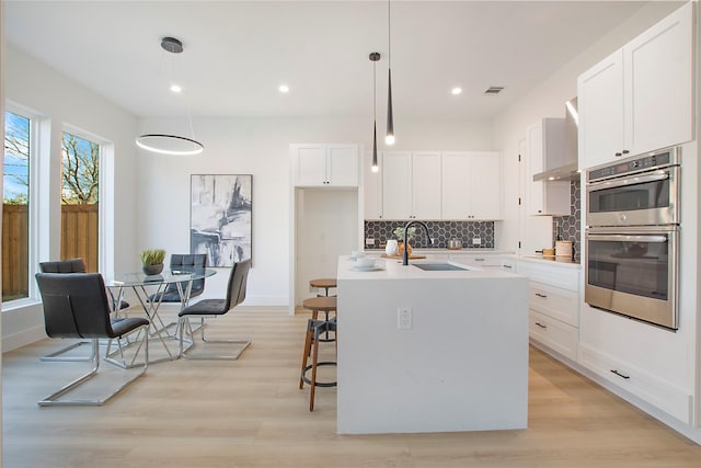 kitchen with a kitchen island with sink, white cabinets, sink, decorative light fixtures, and stainless steel double oven