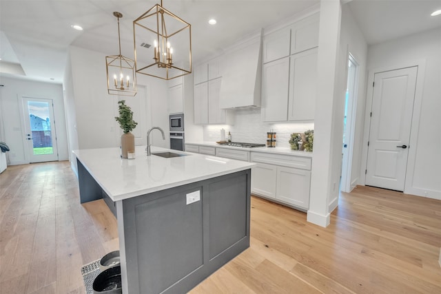 kitchen with custom range hood, light hardwood / wood-style flooring, hanging light fixtures, and an island with sink