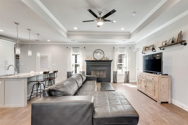 living room featuring sink, light hardwood / wood-style flooring, ceiling fan, ornamental molding, and a tray ceiling