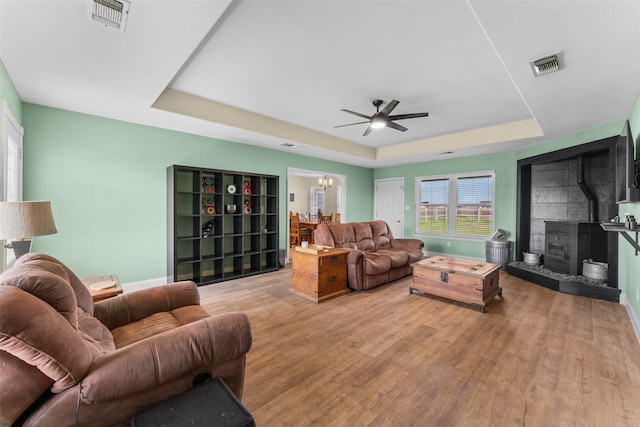 living room with a raised ceiling, a wood stove, wood-type flooring, and ceiling fan with notable chandelier