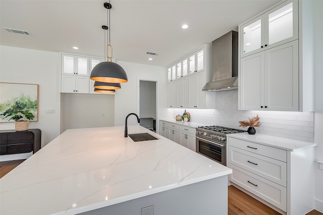 kitchen with a center island with sink, white cabinets, wall chimney range hood, sink, and hanging light fixtures