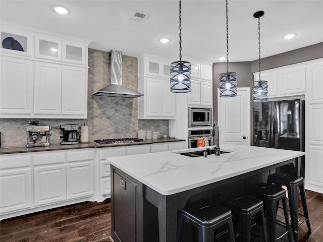 kitchen featuring a center island with sink, dark stone counters, wall chimney range hood, and appliances with stainless steel finishes