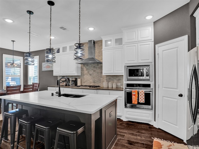 kitchen with sink, stainless steel appliances, wall chimney range hood, a kitchen island with sink, and white cabinets