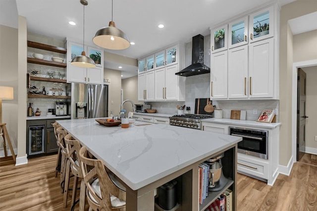 kitchen with white cabinets, an island with sink, stainless steel appliances, and wall chimney range hood