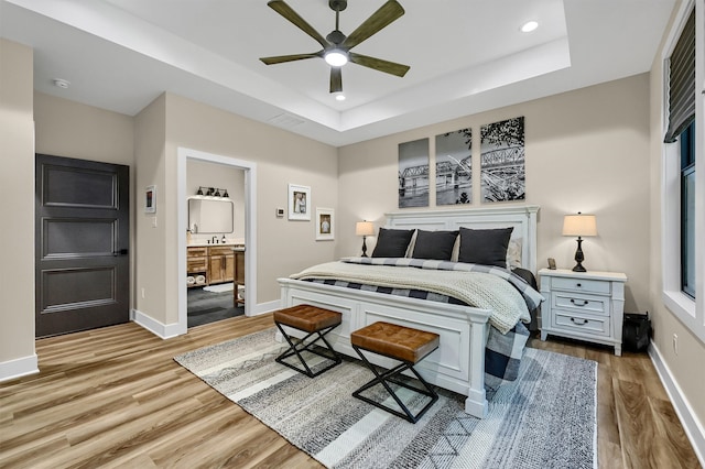 bedroom featuring a tray ceiling, ensuite bath, ceiling fan, and light wood-type flooring