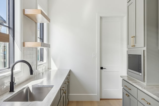 kitchen with stainless steel microwave, light stone countertops, gray cabinets, open shelves, and a sink