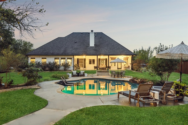 back of house at dusk with an outdoor pool, a chimney, a lawn, a shingled roof, and a patio area