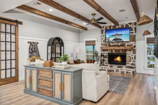 living room with a stone fireplace, ceiling fan, a barn door, beam ceiling, and wood-type flooring