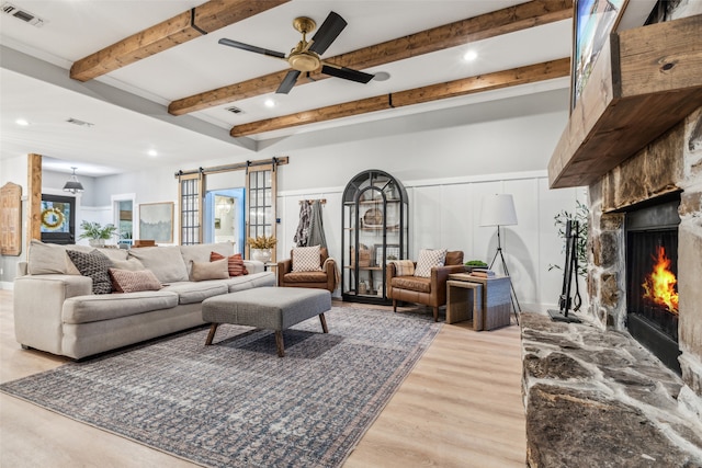 living room featuring ceiling fan, a barn door, light hardwood / wood-style flooring, beamed ceiling, and a fireplace