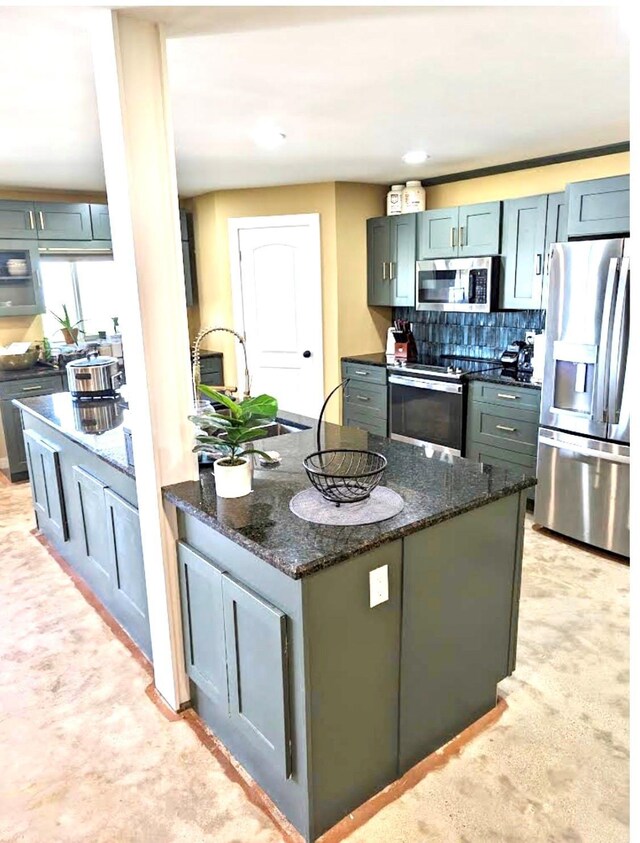 kitchen featuring stainless steel appliances, backsplash, dark stone countertops, an island with sink, and light carpet