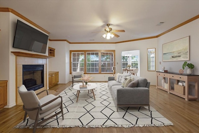 living room featuring ceiling fan with notable chandelier, ornamental molding, a tiled fireplace, and light hardwood / wood-style floors