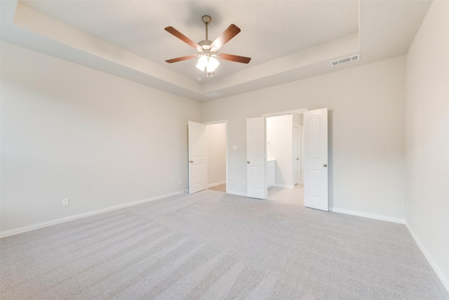 unfurnished bedroom with ceiling fan, a tray ceiling, light colored carpet, and ensuite bath