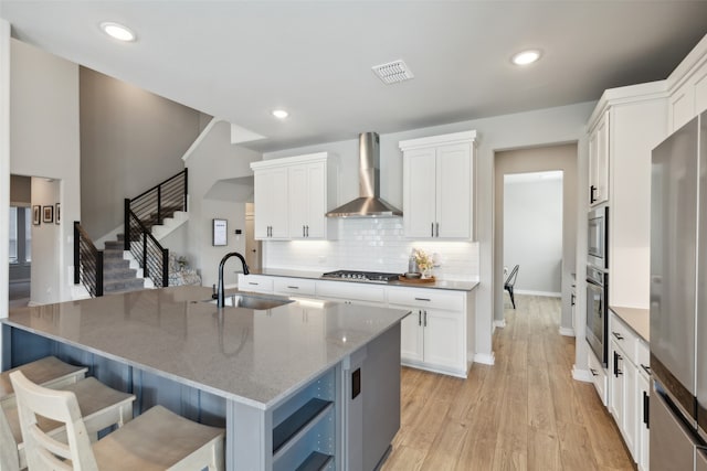 kitchen with white cabinets, wall chimney range hood, sink, and an island with sink