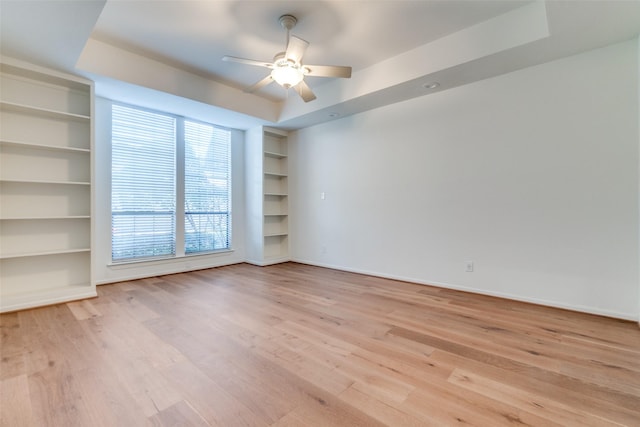 empty room featuring light wood finished floors, a tray ceiling, a ceiling fan, and built in features