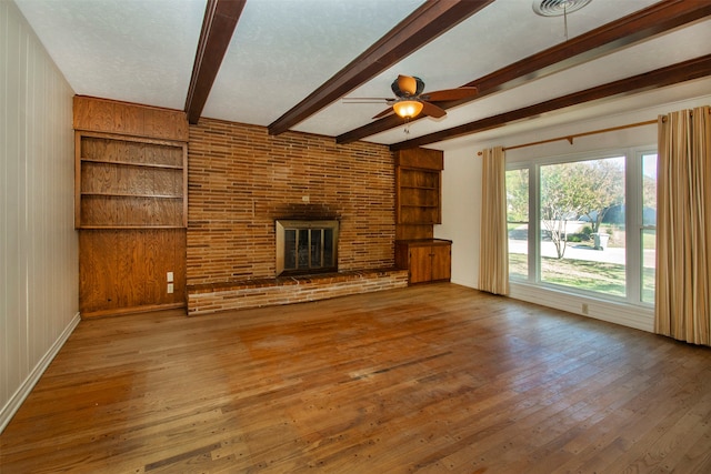 unfurnished living room featuring a brick fireplace, ceiling fan, wooden walls, hardwood / wood-style flooring, and beamed ceiling