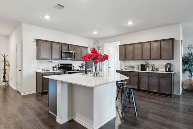 kitchen featuring dark brown cabinetry, dark wood-type flooring, stainless steel appliances, a breakfast bar area, and a kitchen island with sink