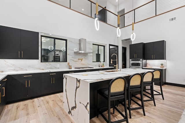 kitchen featuring wall chimney exhaust hood, a towering ceiling, light hardwood / wood-style floors, a center island with sink, and appliances with stainless steel finishes