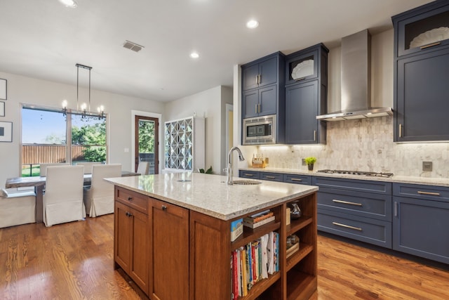 kitchen with light stone countertops, sink, wall chimney range hood, an island with sink, and decorative light fixtures