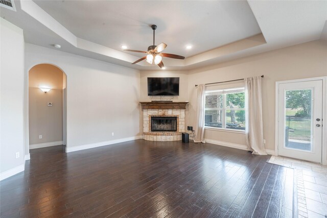 unfurnished living room with ceiling fan, a stone fireplace, dark wood-type flooring, and a tray ceiling