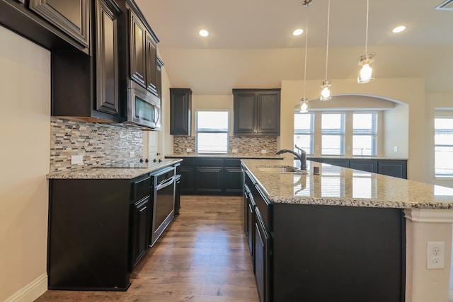 kitchen with hardwood / wood-style floors, backsplash, light stone countertops, an island with sink, and stainless steel appliances