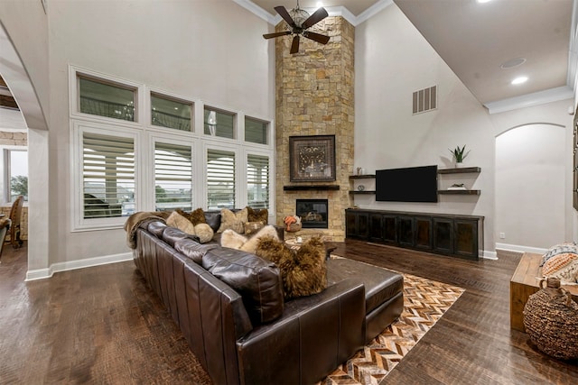 living room featuring a towering ceiling, ceiling fan, dark wood-type flooring, crown molding, and a stone fireplace