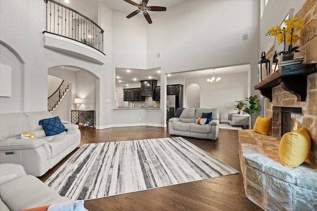 living area featuring stairway, dark wood-style floors, arched walkways, a stone fireplace, and ceiling fan with notable chandelier