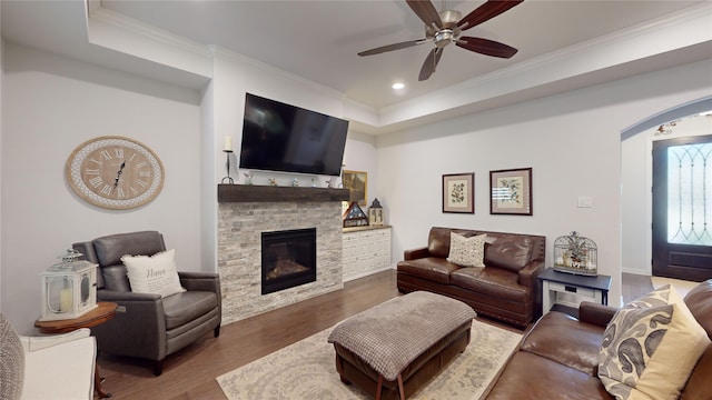 living room featuring a stone fireplace, ceiling fan, dark wood-type flooring, and ornamental molding