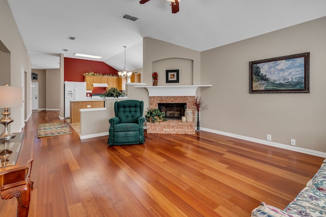 living room featuring hardwood / wood-style floors, ceiling fan with notable chandelier, vaulted ceiling, and a brick fireplace