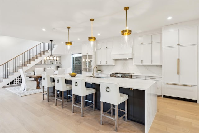 kitchen with sink, paneled built in refrigerator, premium range hood, hanging light fixtures, and a large island with sink