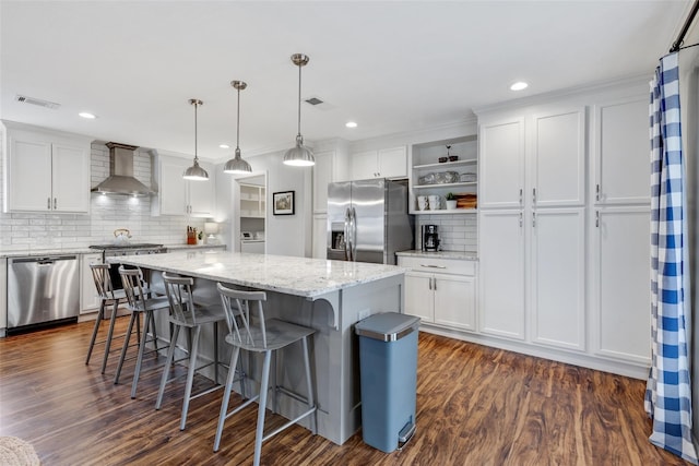 kitchen featuring pendant lighting, wall chimney exhaust hood, a kitchen island, white cabinetry, and stainless steel appliances