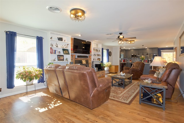 living room with built in shelves, ceiling fan, light hardwood / wood-style flooring, a fireplace, and ornamental molding