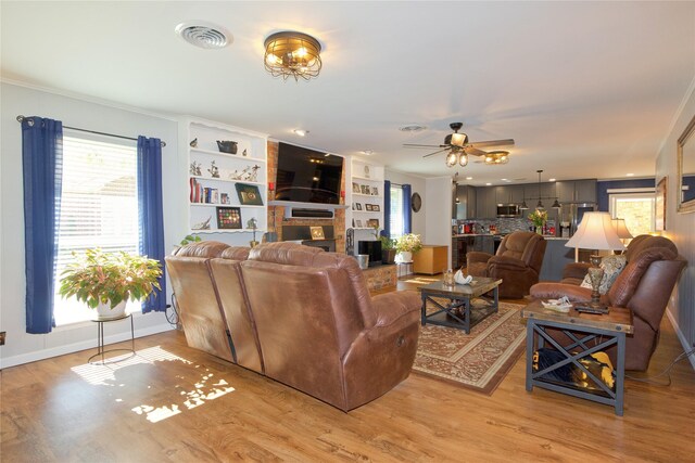 living room with built in shelves, ceiling fan, light hardwood / wood-style flooring, a fireplace, and ornamental molding