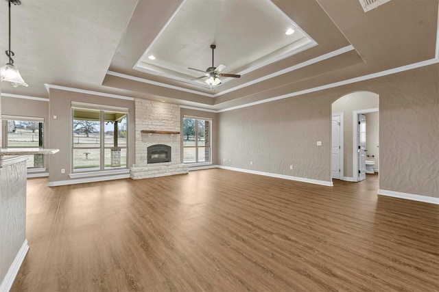 unfurnished living room with a stone fireplace, a raised ceiling, ceiling fan, ornamental molding, and wood-type flooring