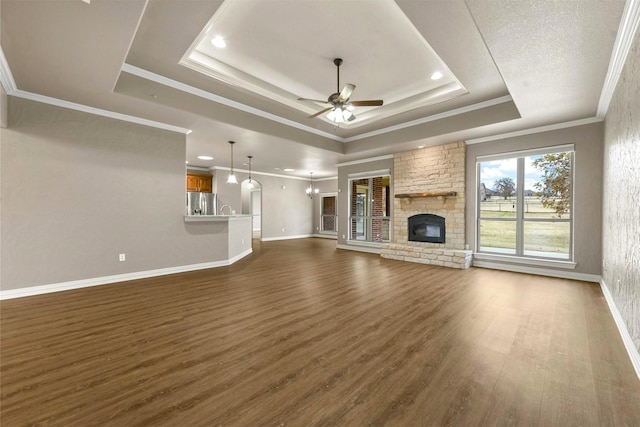 unfurnished living room featuring dark wood-type flooring, a stone fireplace, a tray ceiling, ceiling fan with notable chandelier, and ornamental molding