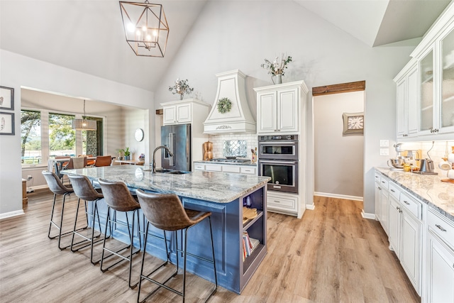 kitchen featuring light stone countertops, custom range hood, stainless steel appliances, hanging light fixtures, and an island with sink