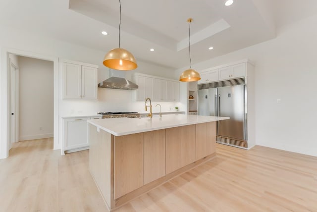 kitchen featuring built in fridge, a raised ceiling, light wood-style floors, and wall chimney range hood