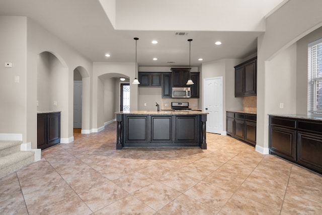 kitchen featuring light stone counters, stainless steel appliances, a kitchen island with sink, sink, and hanging light fixtures