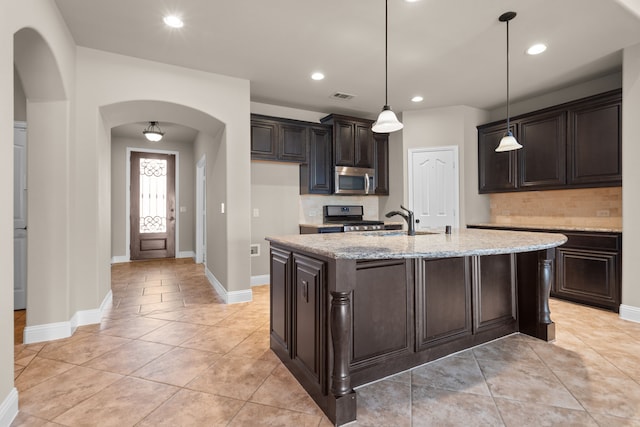 kitchen with a center island with sink, black range with gas stovetop, hanging light fixtures, light stone countertops, and dark brown cabinets