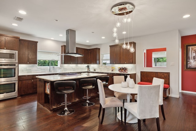 kitchen with dark wood-type flooring, hanging light fixtures, a kitchen island, island range hood, and stainless steel appliances