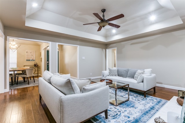 living room featuring ceiling fan with notable chandelier, a raised ceiling, and dark wood-type flooring