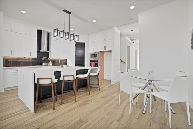 kitchen with wall chimney exhaust hood, light hardwood / wood-style flooring, a center island with sink, white cabinets, and appliances with stainless steel finishes