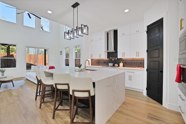 kitchen with wall chimney range hood, plenty of natural light, an island with sink, white cabinets, and light wood-type flooring